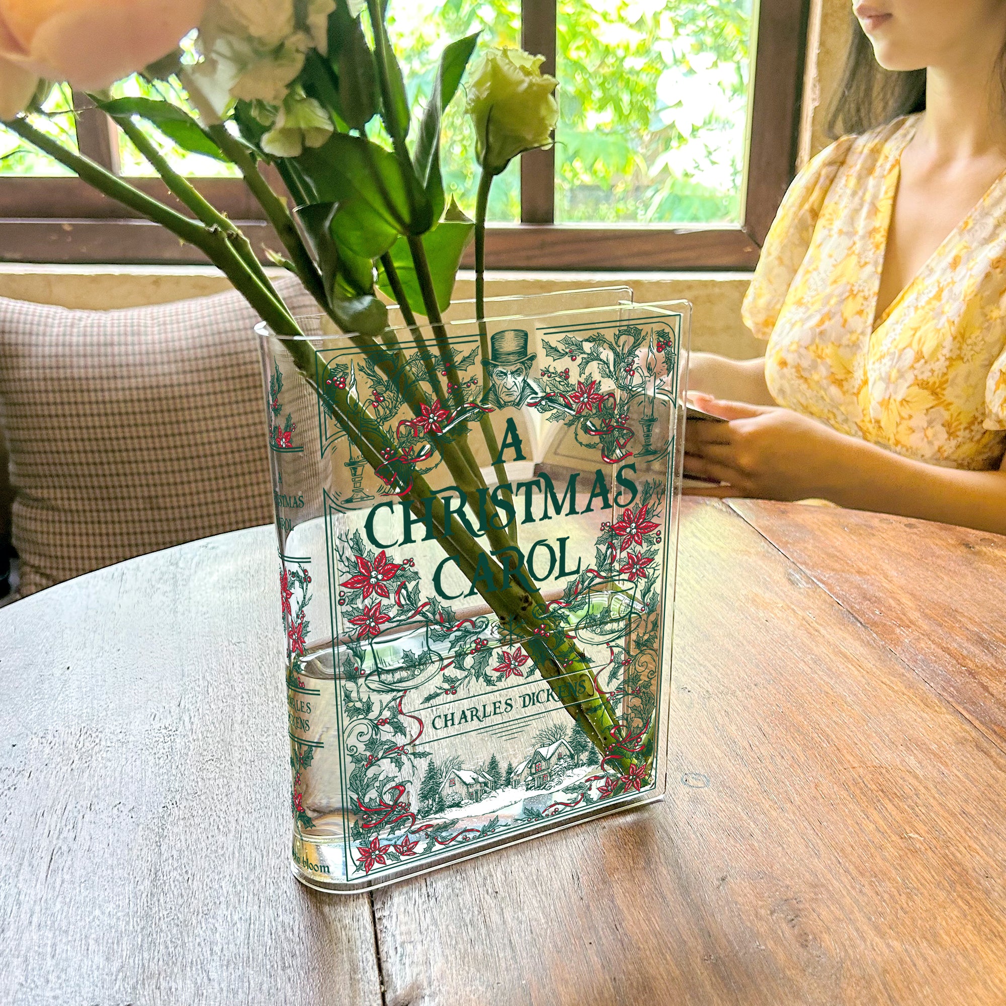 Acrylic book vase with A Christmas Carol cover holding flowers on wooden table near woman