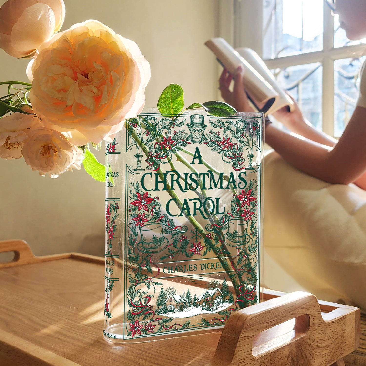 A Christmas Carol acrylic book vase with floral design on a wooden tray, flowers and a person reading by a window.