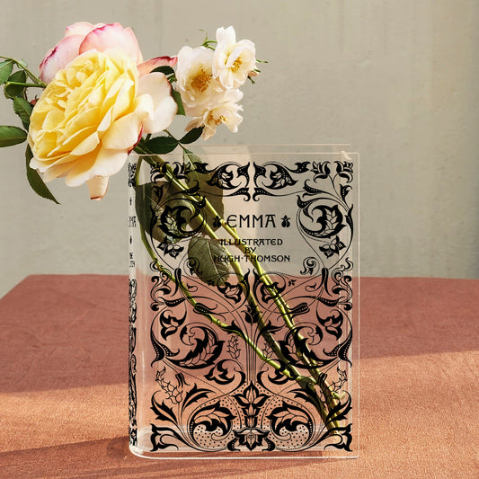 Acrylic book vase with 'Emma' floral design holding yellow and white roses on a pink table.