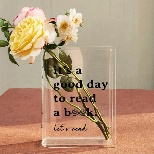 Acrylic book vase with bouquet and black text 'it's a good day to read a book' on table.
