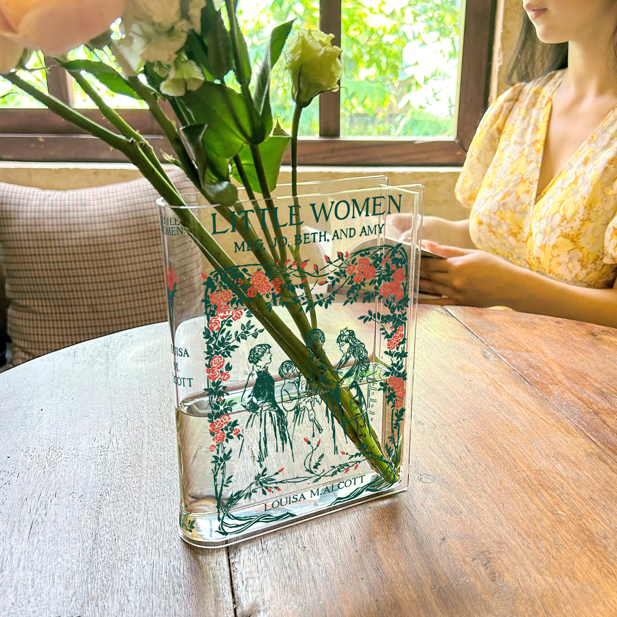 Little Women acrylic book vase with flowers on wood table in sunny cafe with woman in yellow dress