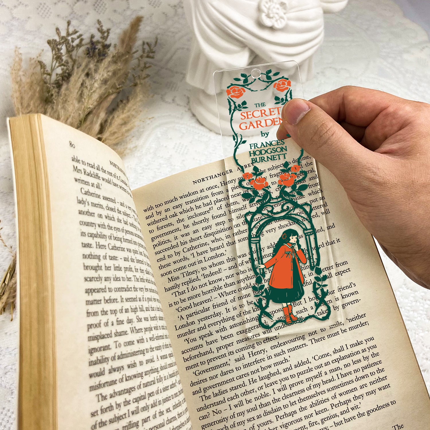 Hand holding The Secret Garden clear acrylic bookmark over open book with floral decor