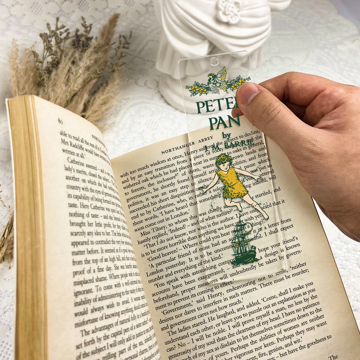 Hand holding Peter Pan clear bookmark over open book on lace table with dried flowers