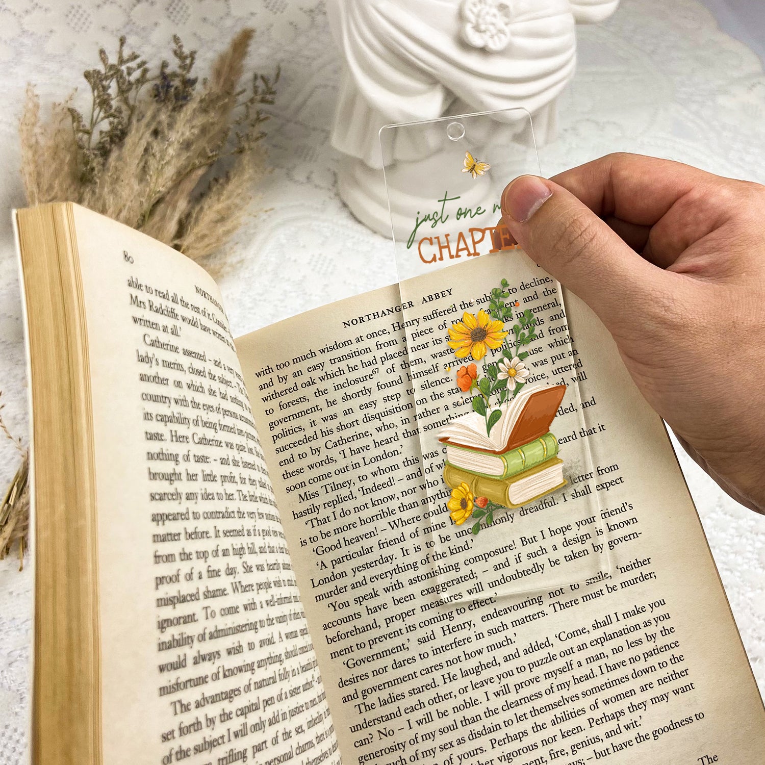 Hand holding clear floral bookmark with books and 'just one more chapter' in open novel