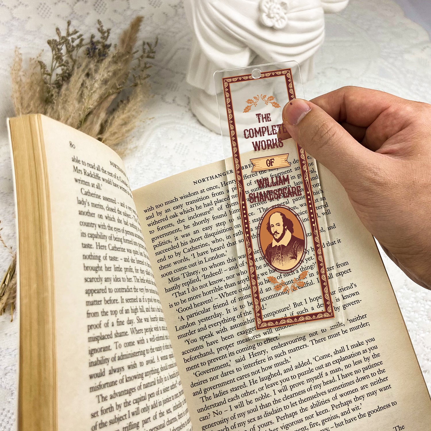 Hand holding Shakespeare bookmark over open book on lace table, dried flowers and white bust nearby.