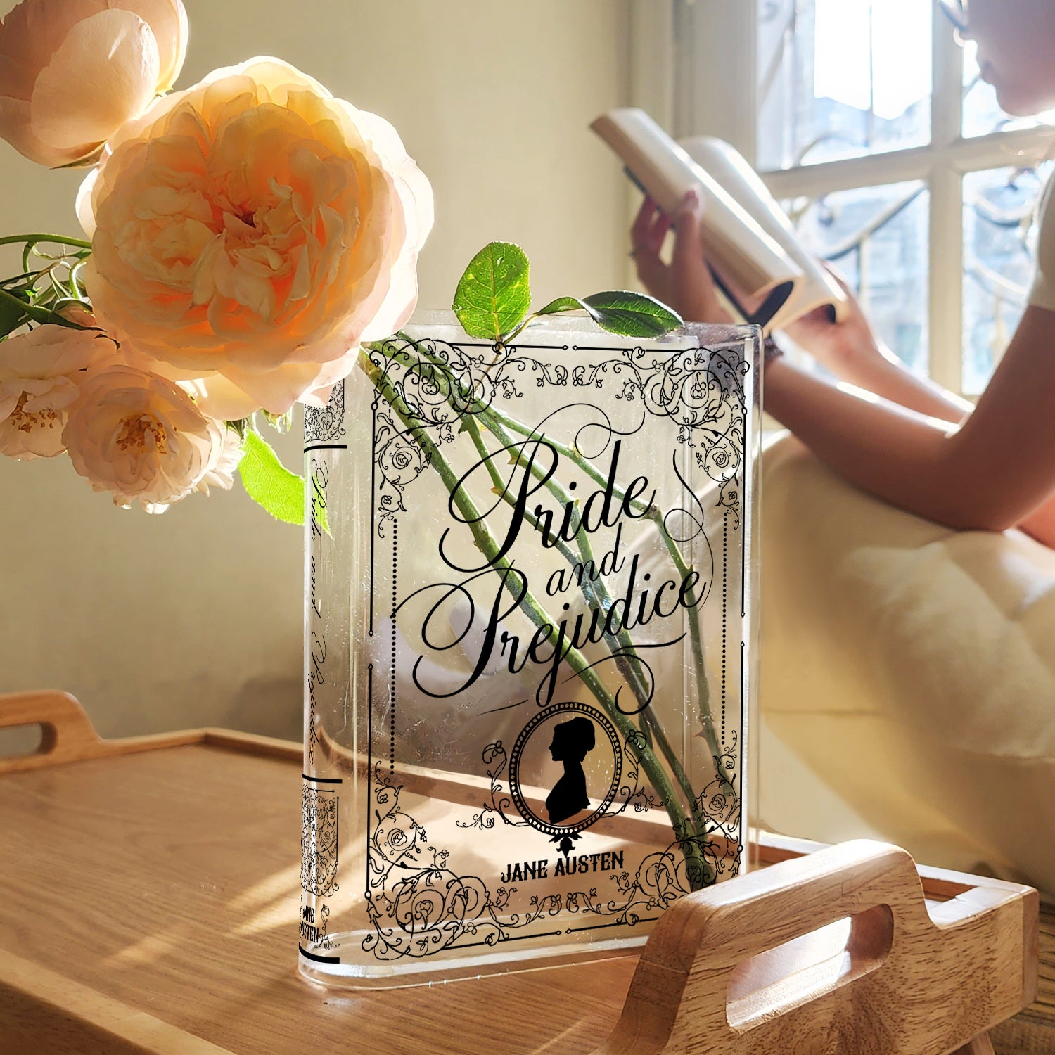 Acrylic book vase with Pride and Prejudice cover, flowers inside, woman reading by window