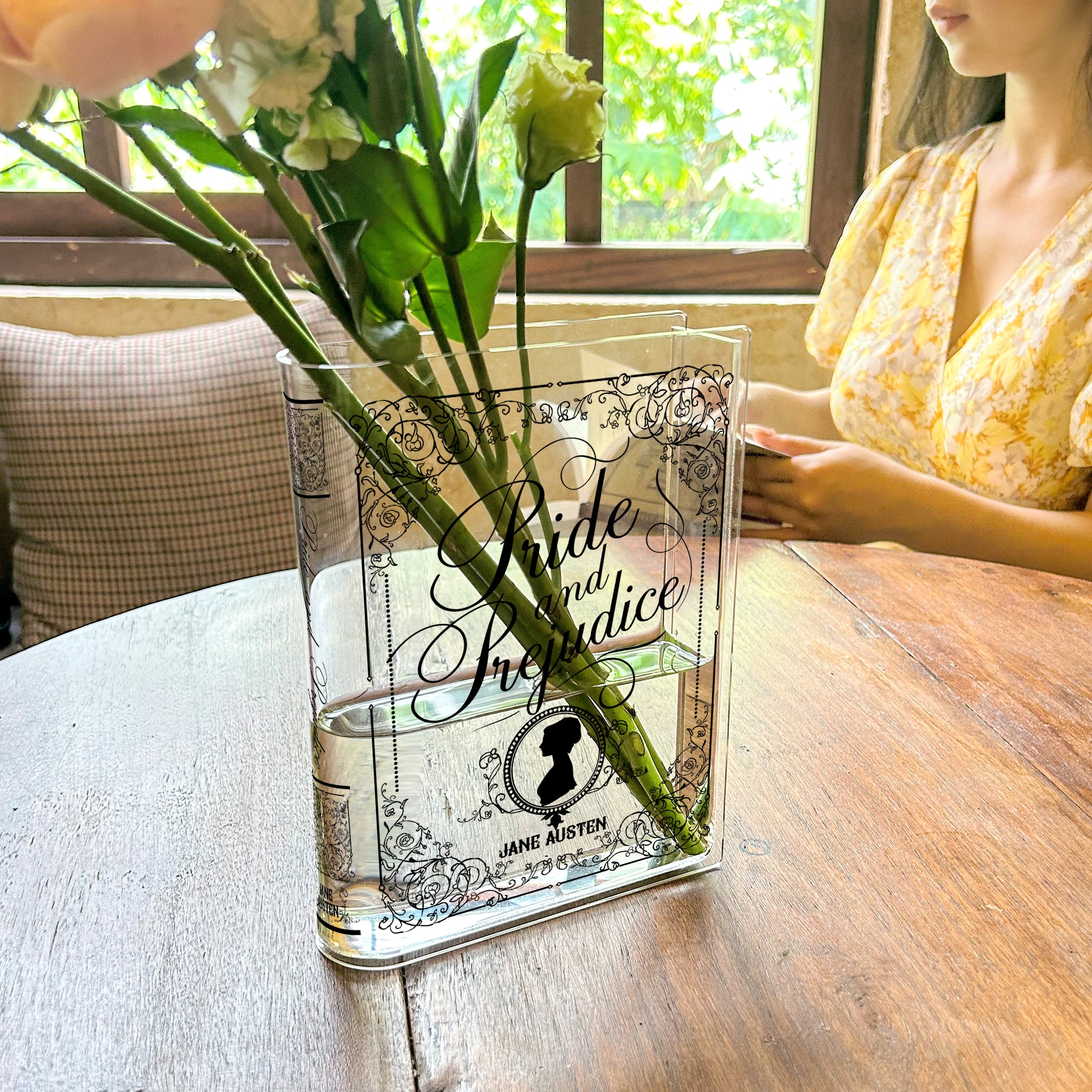 Pride and Prejudice acrylic book vase with flowers on café table, woman in yellow dress nearby