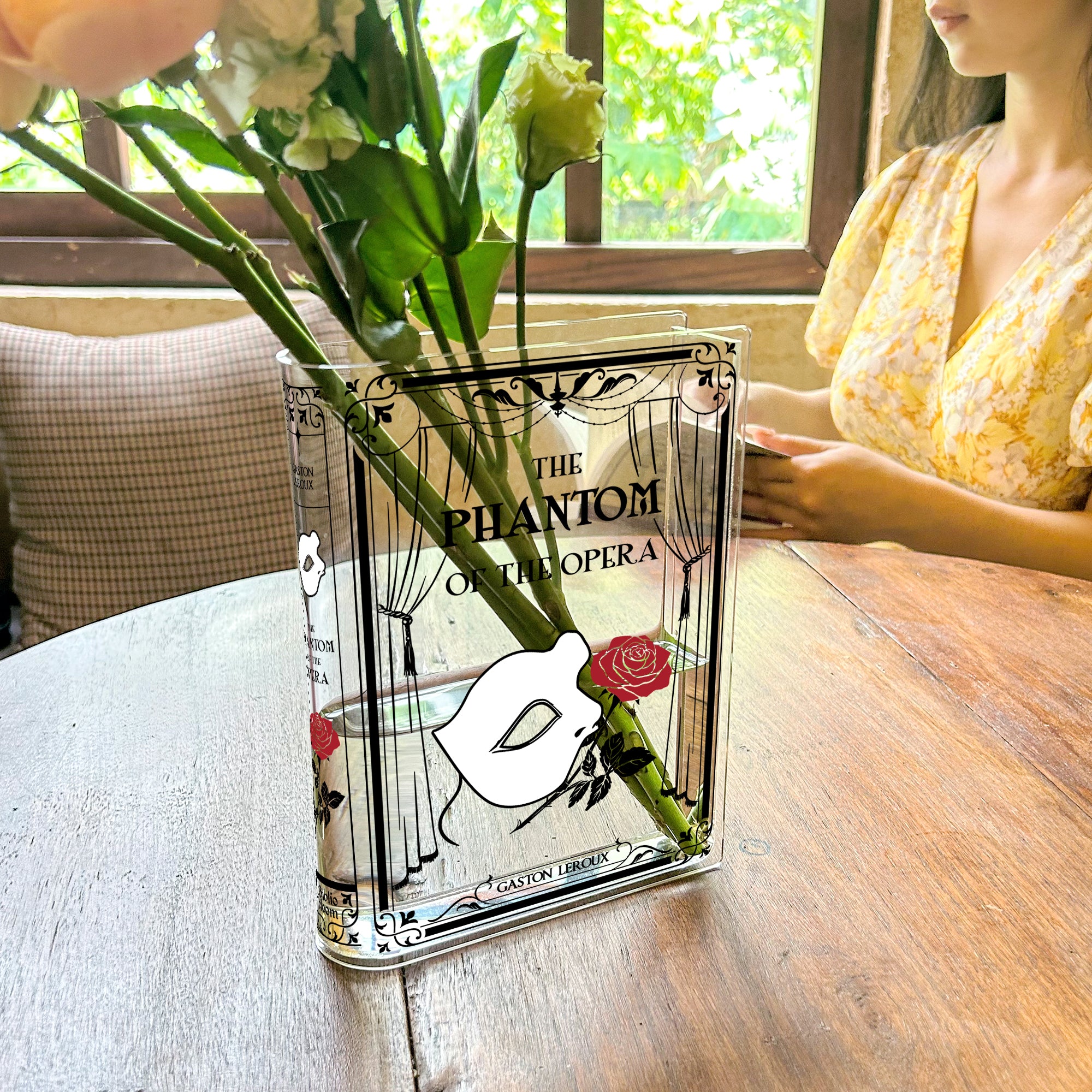 The Phantom of the Opera acrylic book vase with flowers on a wooden table near a window