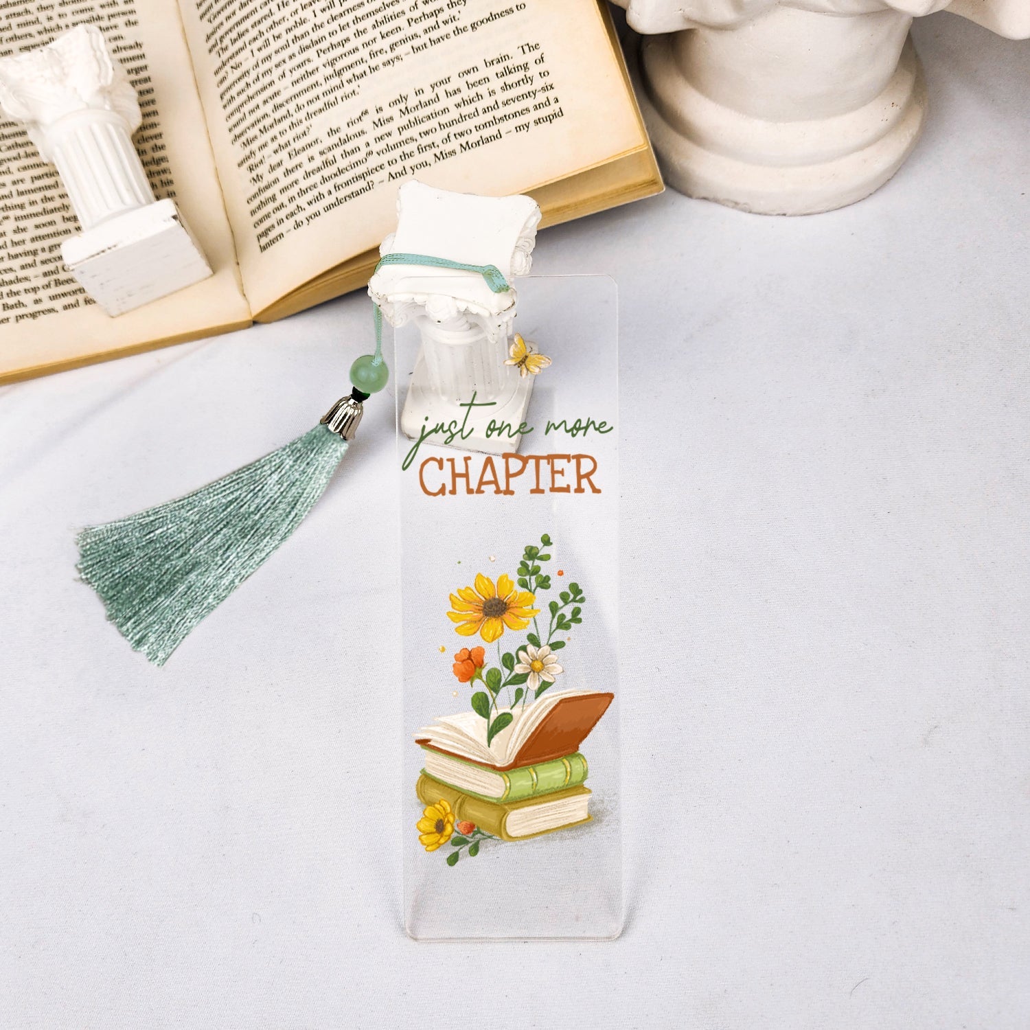 Floral acrylic bookmark with 'just one more chapter' text and green tassel beside open book.