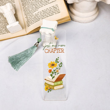 Floral clear bookmark with 'just one more chapter' text, tassel, and books, beside open book.